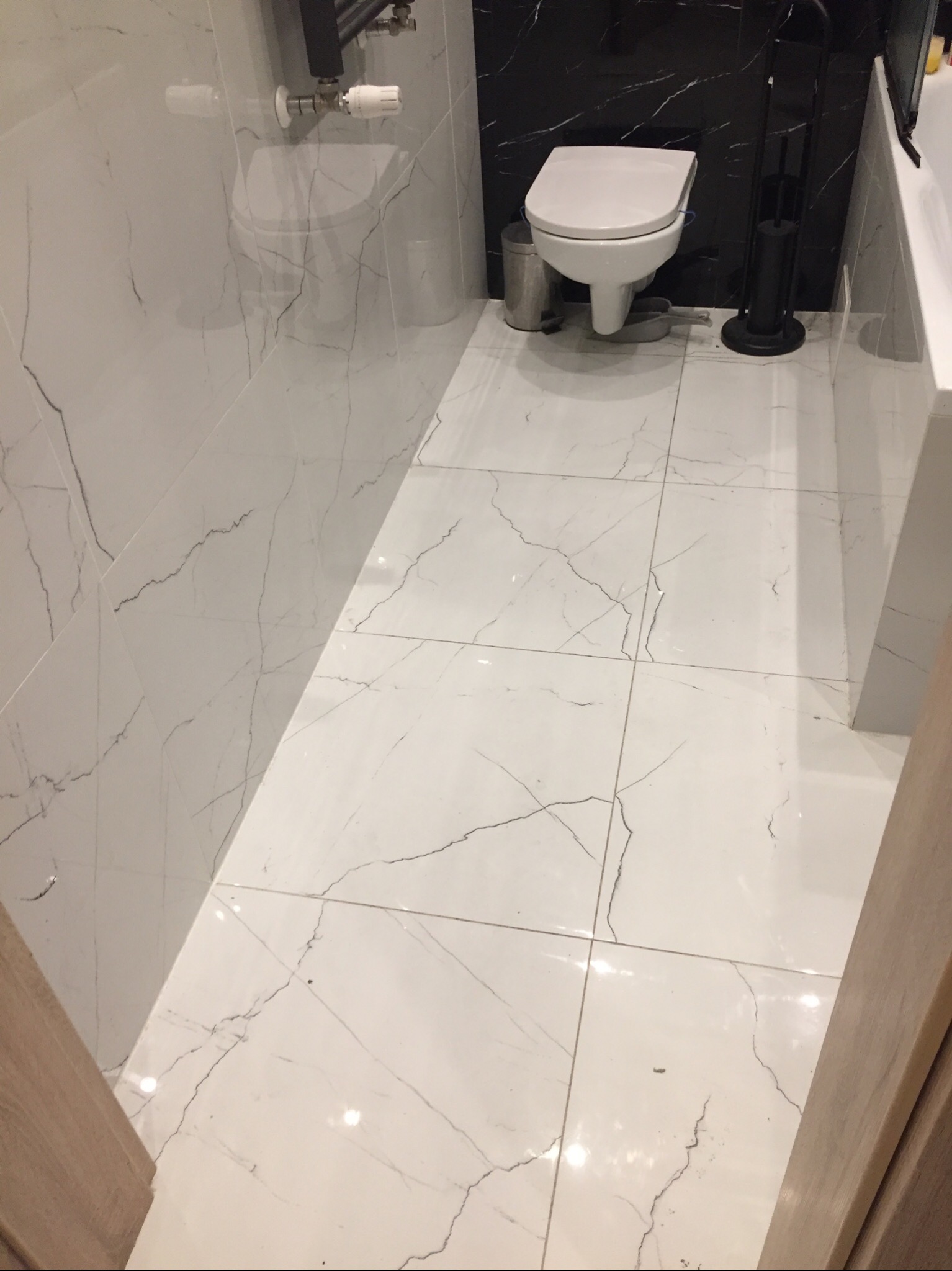 Wykończona łazienka z białymi płytkami imitującymi marmur na podłodze i ścianach, z czarnym akcentem na ścianie za toaletą i akcesoriami łazienkowymi.