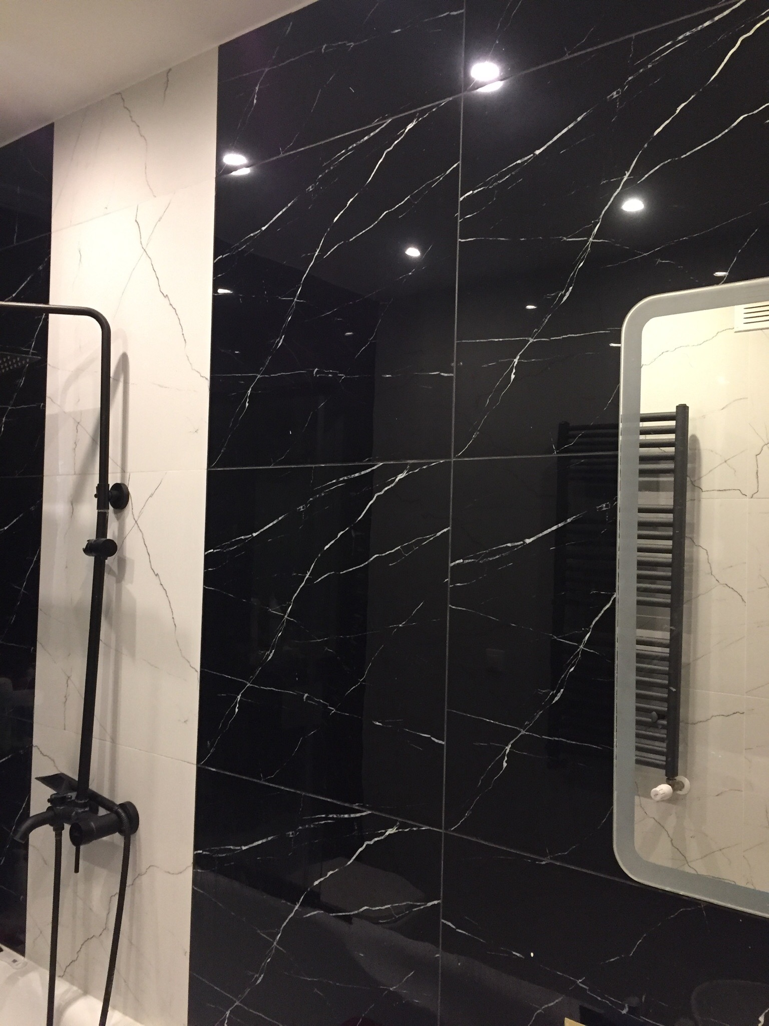 Nowoczesna łazienka z czarno-białymi płytkami imitującymi marmur, czarna armatura prysznicowa i grzejnik drabinkowy odbijający się w lustrze.