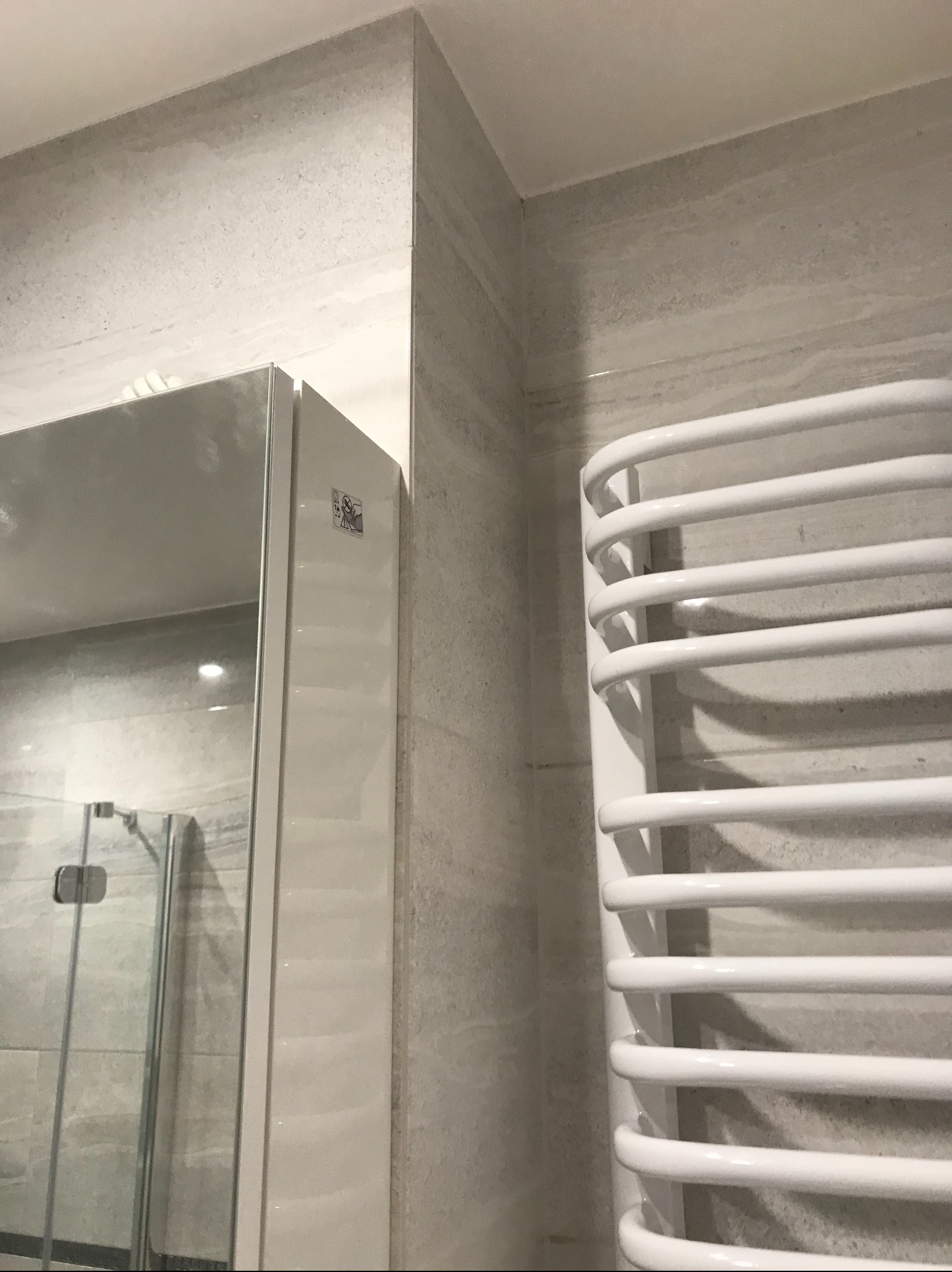 Fragment łazienki z grzejnikiem drabinkowym, lustrem z kabiną prysznicową w odbiciu i płytkami imitującymi kamień na ścianach.