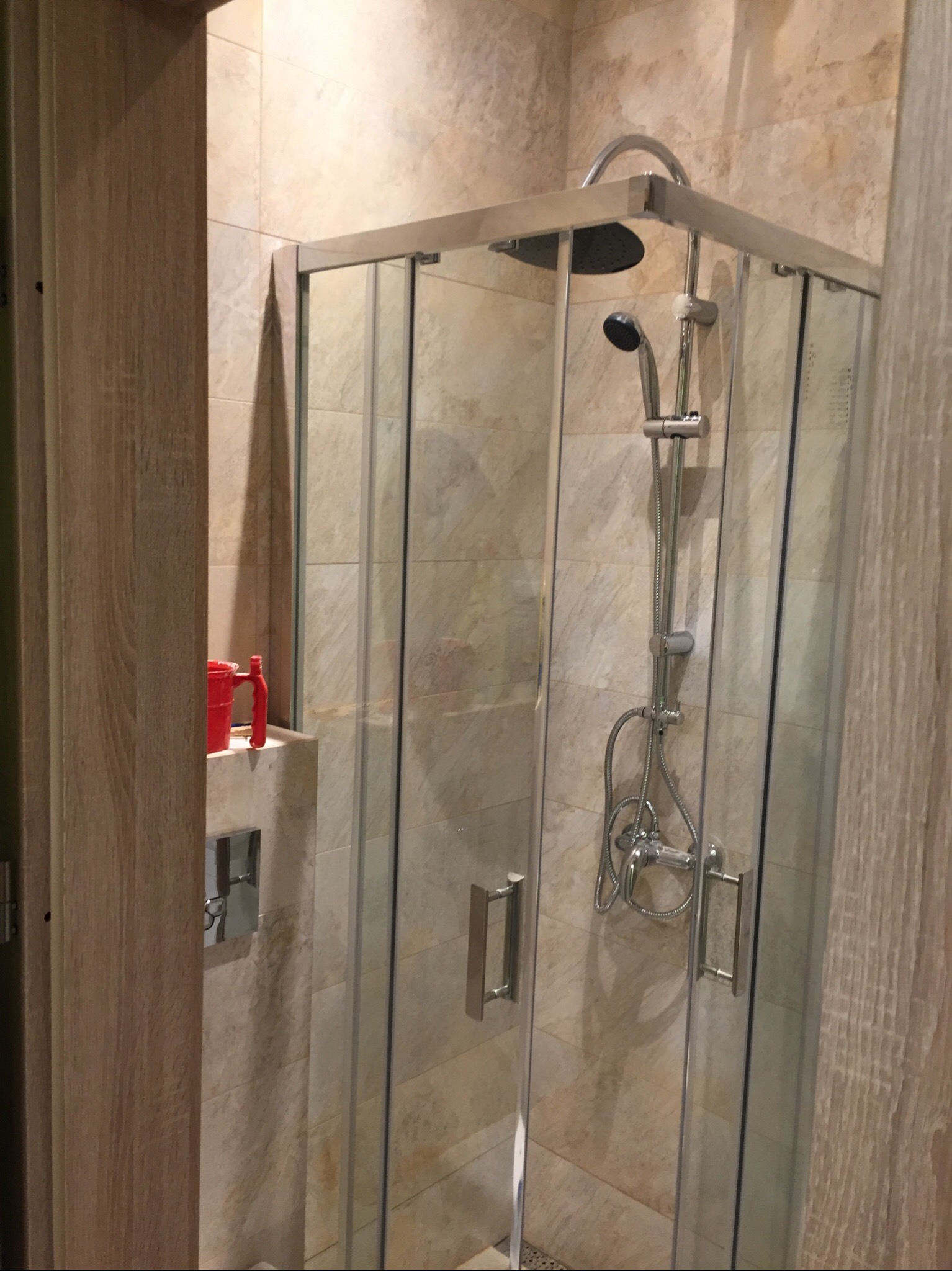 Zamknięta kabina prysznicowa z przesuwnymi drzwiami, wykończona beżowymi płytkami imitującymi kamień, z widoczną baterią prysznicową i deszczownicą oraz czerwonym kubkiem na półce.