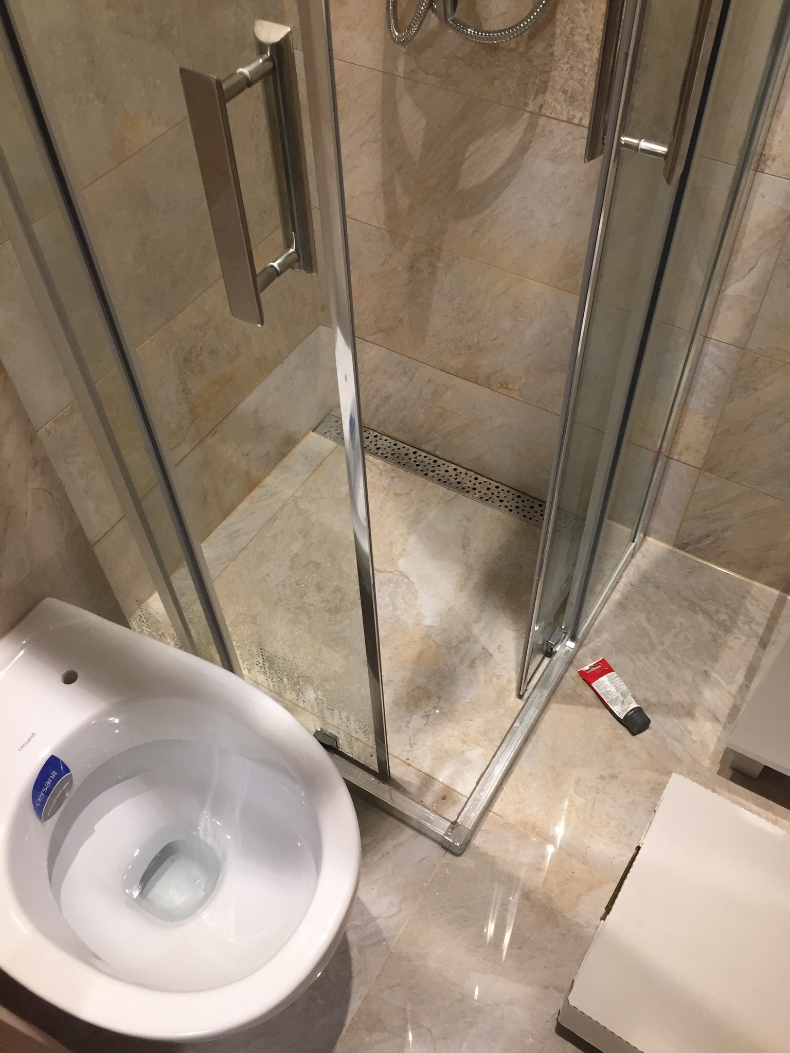 Narożnik łazienki z kabiną prysznicową z chromowanymi okuciami, wiszącą toaletą marki Cersanit i beżowymi płytkami podłogowymi; tuba uszczelniacza silikonowego leży przy krawędzi brodzika.