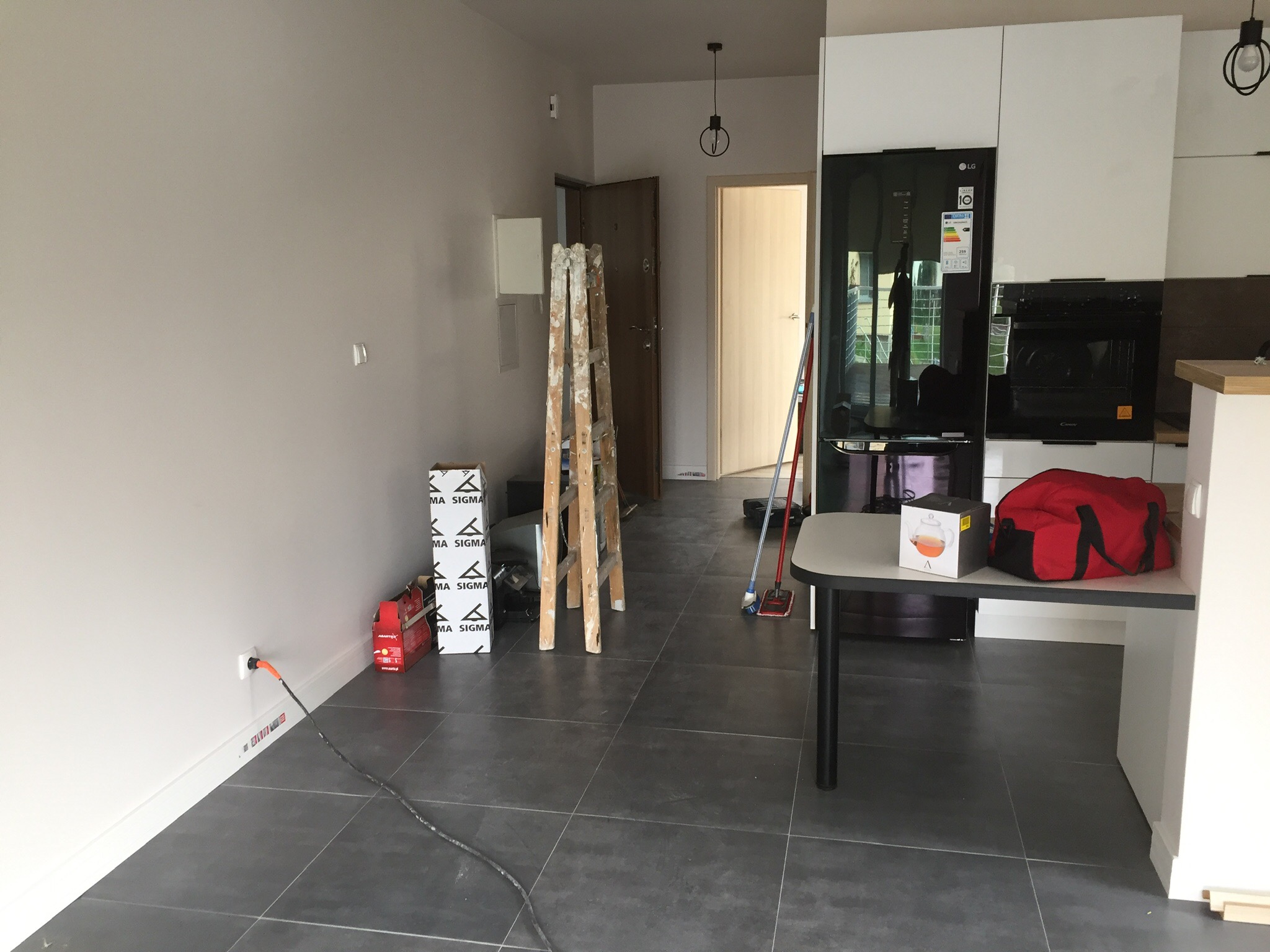 Wnętrze mieszkania w trakcie remontu: drabina malarska, kartony z materiałami budowlanymi, podłoga z szarych płytek, widoczne elementy kuchni.