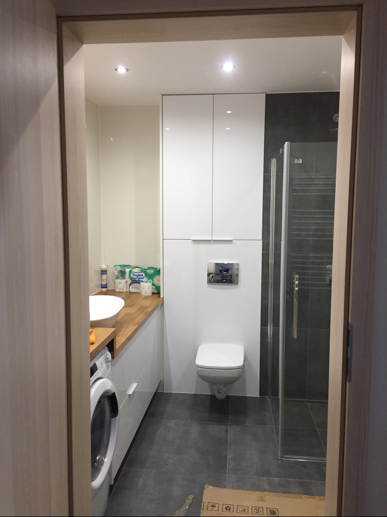Nowoczesna łazienka z pralką, umywalką na drewnianym blacie, kabiną prysznicową z ciemnymi płytkami i podwieszaną toaletą z chromowanym przyciskiem spłukiwania, widziana zza progu.
