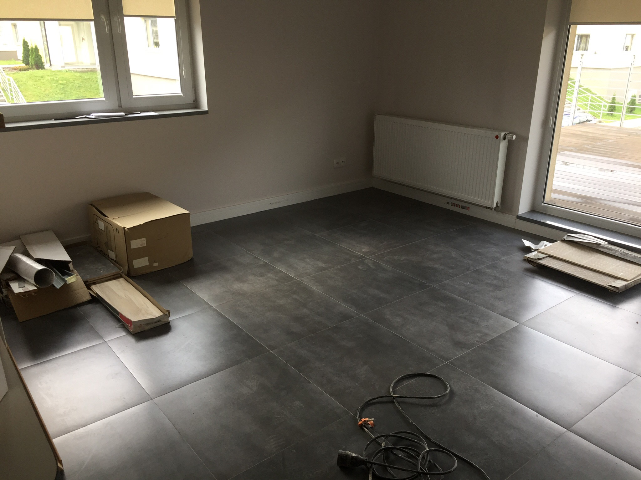 Wnętrze pokoju z ułożonymi ciemnoszarymi płytkami podłogowymi, widoczne kartony, grzejnik i kabel zasilający na podłodze, okna z widokiem na zewnątrz.