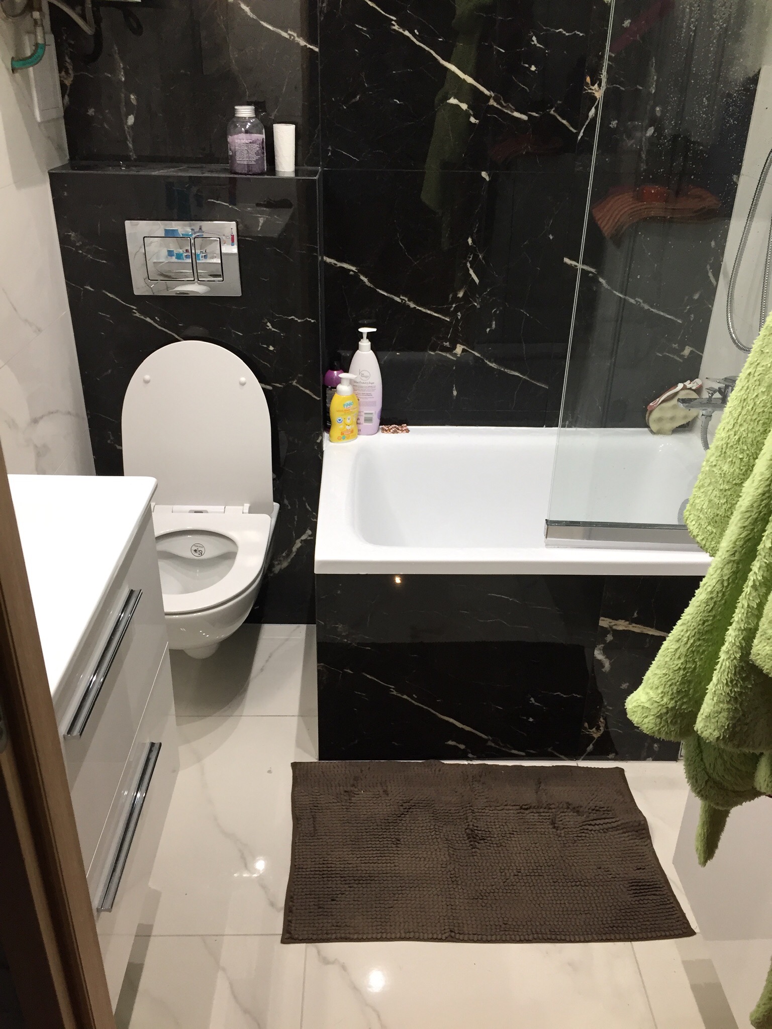 Nowoczesna łazienka z czarnymi marmurowymi płytkami, białą armaturą, wiszącą toaletą z podtynkową spłuczką, białą szafką z szufladami i brązowym dywanikiem.
