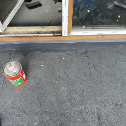 DACHY RYBKA Karol Rybka - Widok z góry na fragment dachu z papy z zamontowanym oknem dachowym z brązowym obramowaniem, obok puszka z farbą i pędzel.