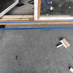 DACHY RYBKA Karol Rybka - Montaż okna dachowego na pokryciu bitumicznym, zabezpieczonym niebieską taśmą malarską, widoczny pędzel, nóż tapicerski i miarka składana.