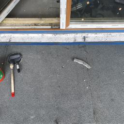 DACHY RYBKA Karol Rybka - Widok z góry na fragment dachu z oknem dachowym, gdzie widoczna jest świeżo położona warstwa hydroizolacyjna wokół okna, obok puszka z uszczelniaczem, pędzel, nóż do cięcia papy i taśma malarska.