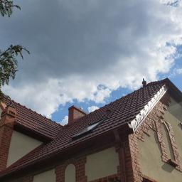 DACHY RYBKA Karol Rybka - Widok z dołu na dwuspadowy dach z brązowej dachówki ceramicznej z oknem połaciowym, kominem i systemem rynnowym, na tle błękitnego nieba z chmurami.