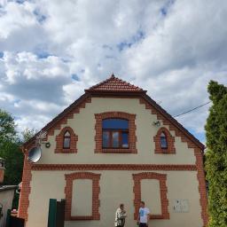 DACHY RYBKA Karol Rybka - Dwupiętrowy dom z czerwonym dachem i ceglanymi zdobieniami wokół okien i na rogach, z anteną satelitarną i dwoma osobami stojącymi przed budynkiem.