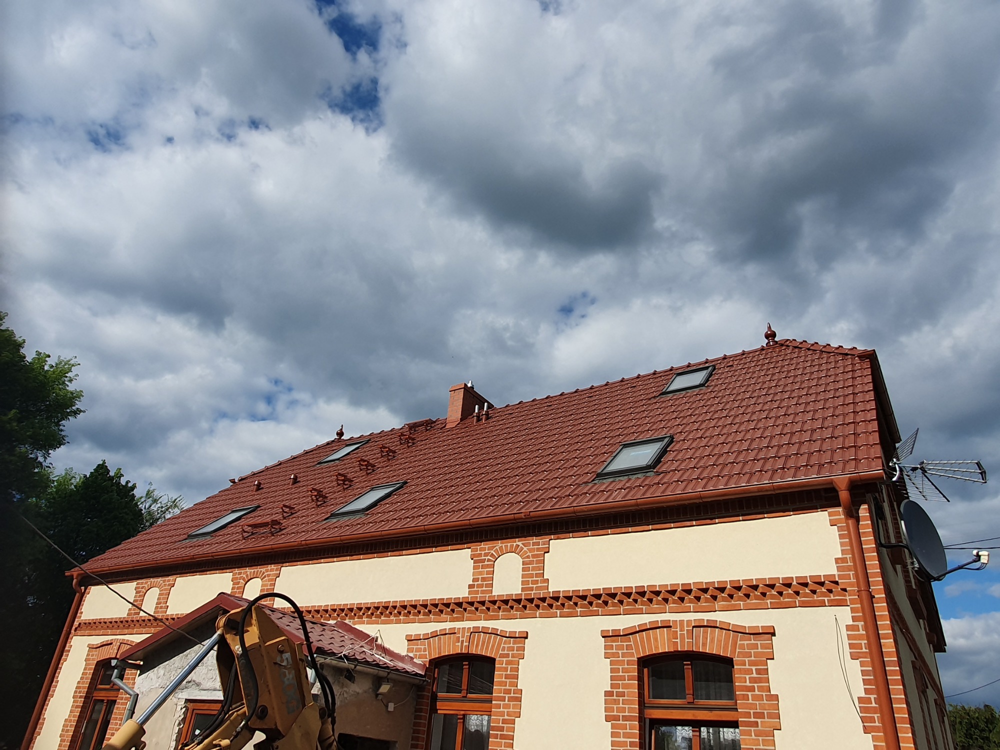 Dom z nowym, czerwonym dachem z dachówki ceramicznej i oknami dachowymi, widoczny fragment koparki.