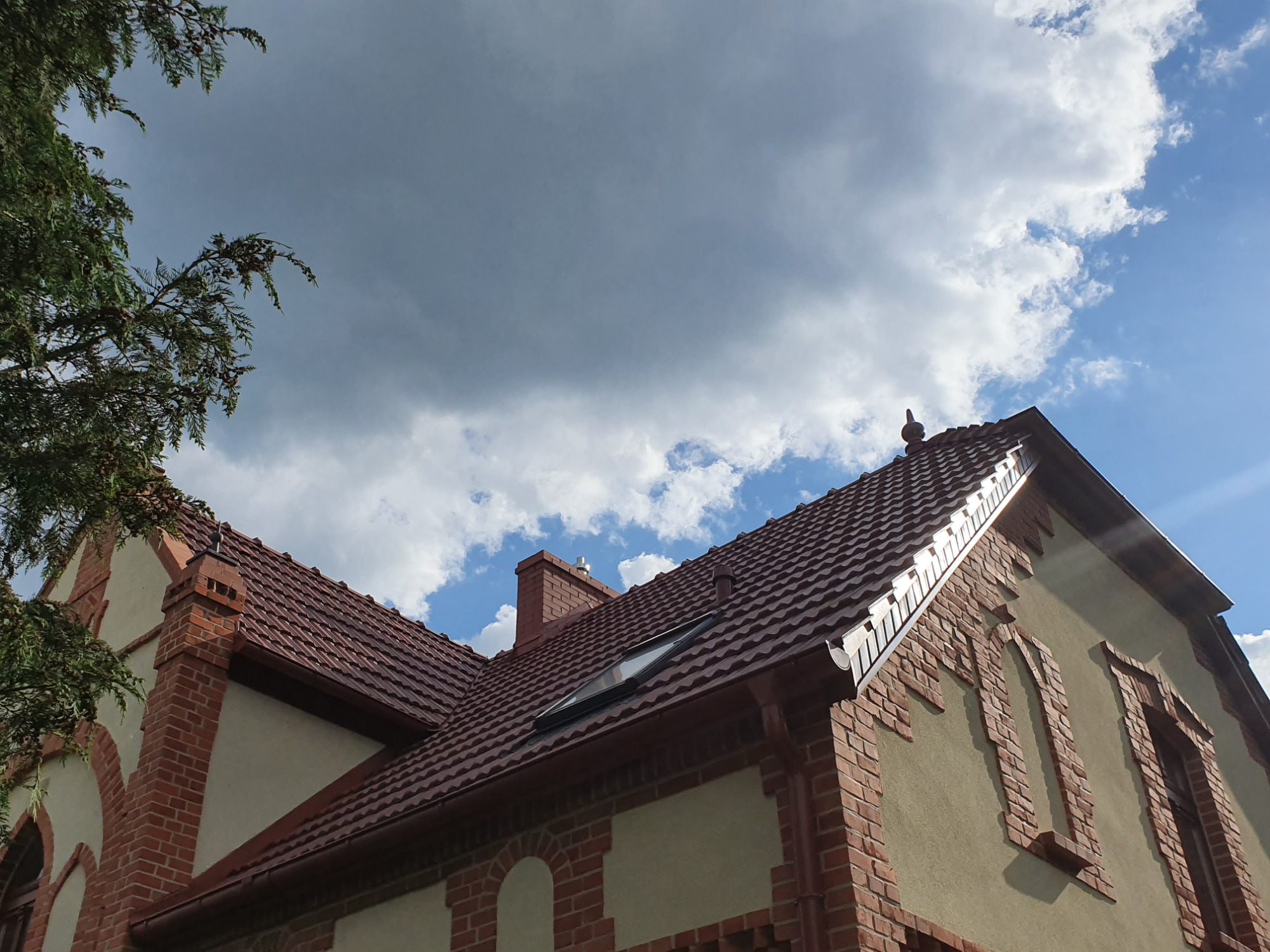 Widok z dołu na dwuspadowy dach z brązowej dachówki ceramicznej z oknem połaciowym, kominem i systemem rynnowym, na tle błękitnego nieba z chmurami.