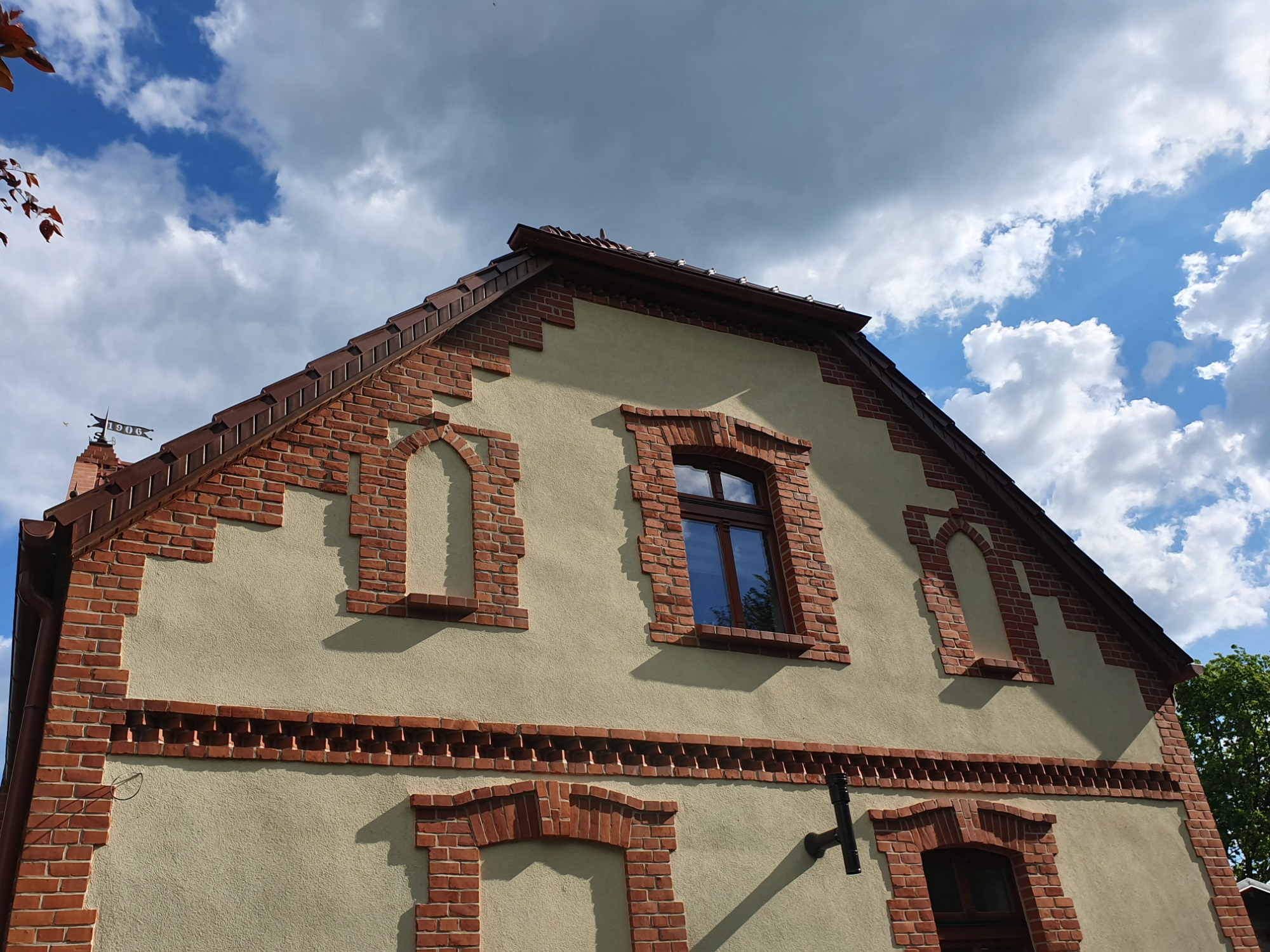 Szczyt budynku z beżową elewacją, obramowany czerwoną cegłą, z brązowym dachem i ozdobną chorągiewką na szczycie, na tle błękitnego nieba z białymi chmurami.