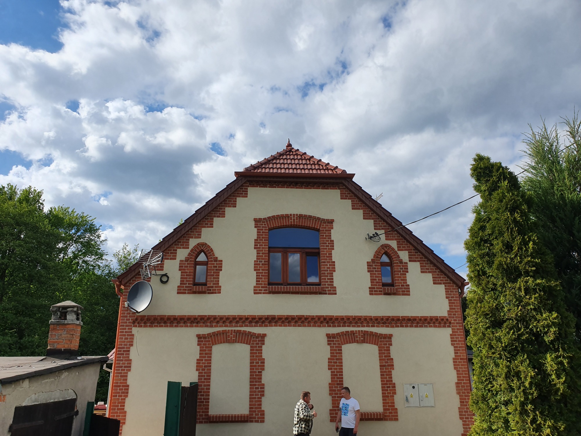 Dwupiętrowy dom z czerwonym dachem i ceglanymi zdobieniami wokół okien i na rogach, z anteną satelitarną i dwoma osobami stojącymi przed budynkiem.