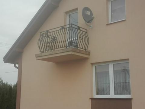 Balkon z czarną, ozdobną balustradą na tle beżowej elewacji budynku z oknami i anteną satelitarną.