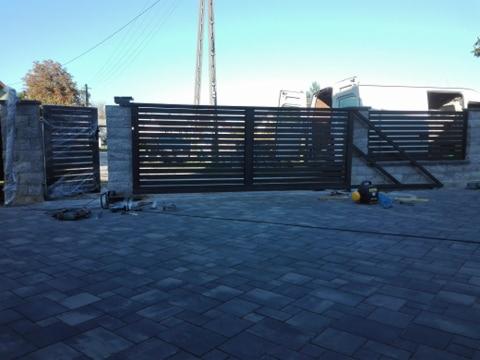 Montaż przesuwnej bramy posesyjnej z poziomych profili, widoczne narzędzia na kostce brukowej, częściowo zasłonięty biały samochód dostawczy w tle.