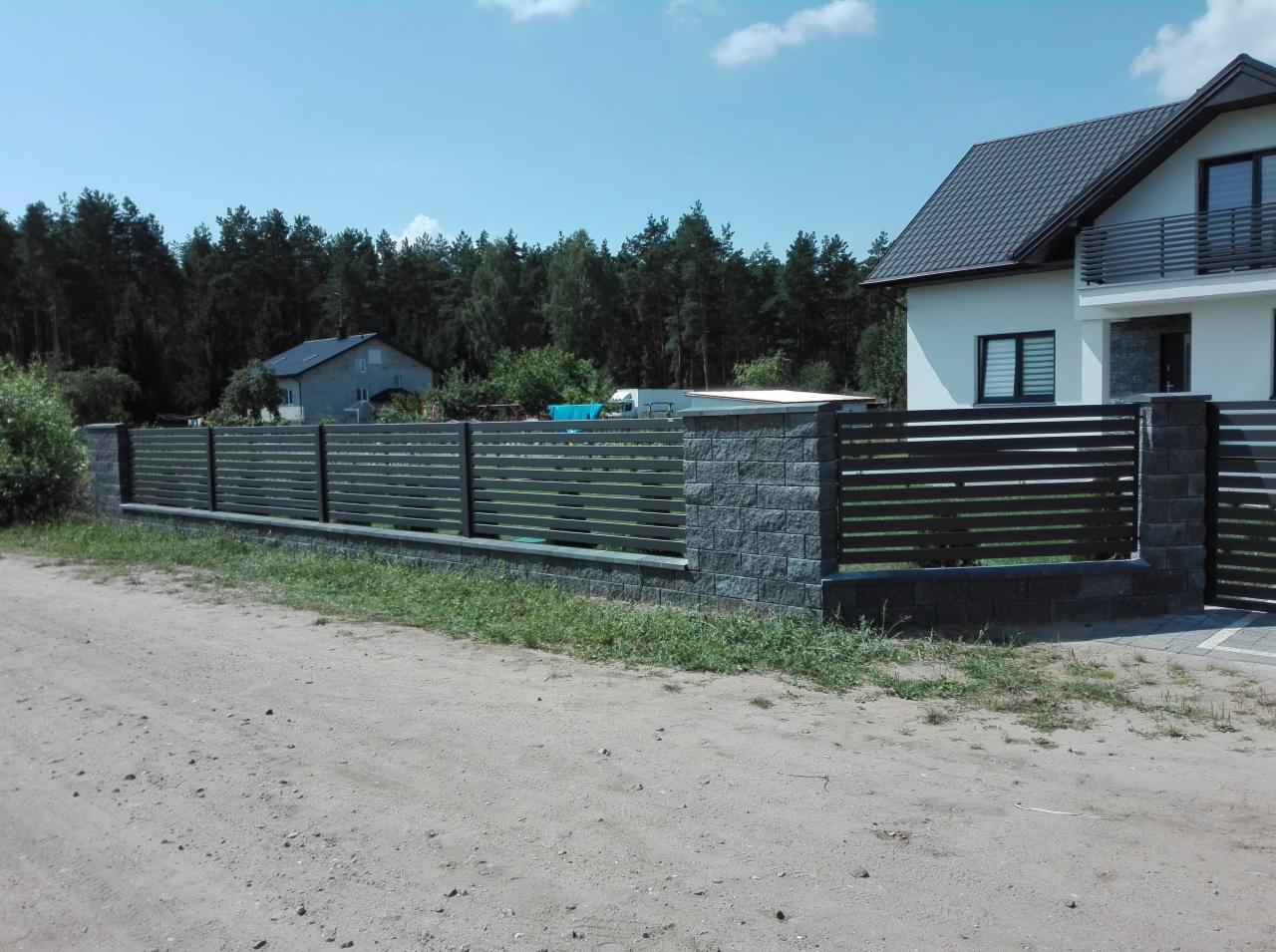 Nowoczesna, pozioma balustrada z szarych, betonowych słupków i ciemnych, metalowych paneli, oddzielająca posesję od piaszczystej drogi, z widocznym domem i lasem w tle.