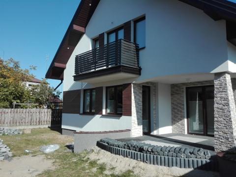 Elewacja domu jednorodzinnego z balkonem z czarną, nowoczesną balustradą, wykończona dekoracyjną okładziną kamienną na filarach wejścia.