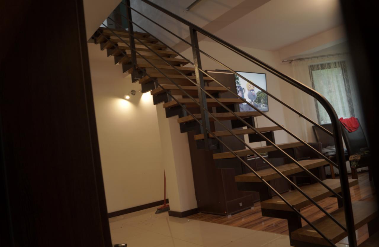 Nowoczesne schody drewniane z czarną, metalową balustradą w jasnym wnętrzu domu z telewizorem w tle.