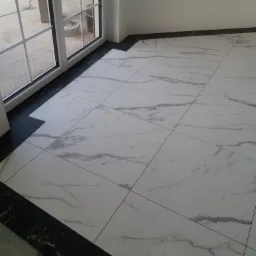 Wykończona podłoga z białych płytek imitujących marmur z czarnym obramowaniem przy oknie balkonowym i ścianie.