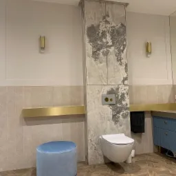 Nowoczesna łazienka z wiszącą toaletą, szafką z marmurowym wzorem, złotymi akcentami i niebieskim pufem.