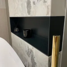 P&oacute;łka wnękowa w łazience wyłożona płytkami imitującymi marmur, z czarnym wnętrzem, na kt&oacute;rej stoją zapach do wnętrz i świeca, widoczny fragment białej wanny i złoty element armatury.