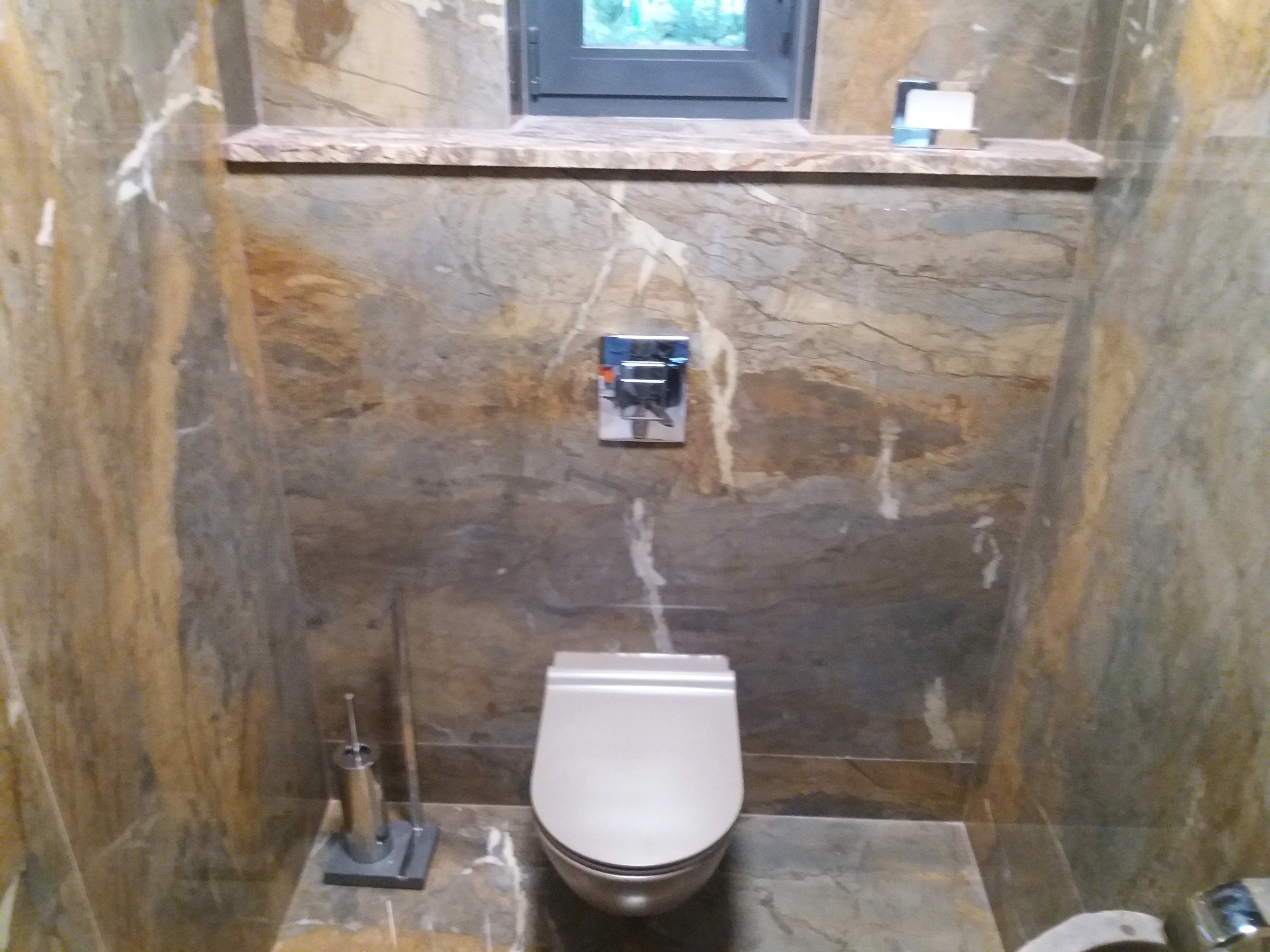 Wykończona toaleta z miską WC w kolorze białym, na ścianach i podłodze płytki imitujące marmur w odcieniach szarości i brązu, widoczny przycisk spłuczki w chromowanej obudowie oraz okno z parapetem.