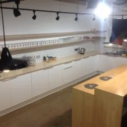 Nowoczesna kuchnia z białymi szafkami, jasnym drewnianym blatem i wyspą z okrągłymi otworami, oświetlona czarnymi reflektorami i wiszącą lampą, z płytkami w kratkę na ścianie.