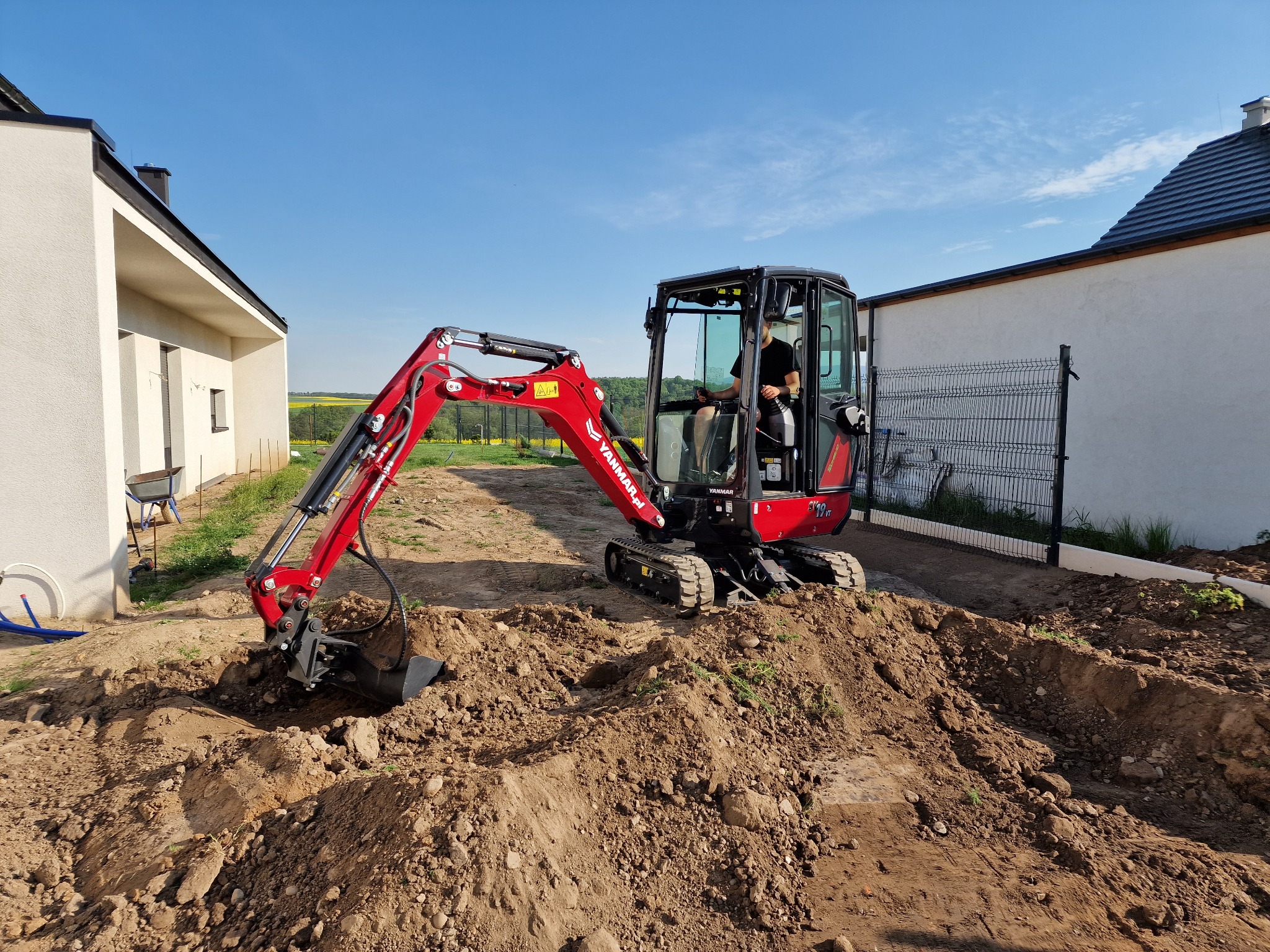 Czerwona mini koparka Yanmar na placu budowy, operator w kabinie, świeżo wykopana ziemia, w tle nowoczesny dom i żółte pole rzepaku.