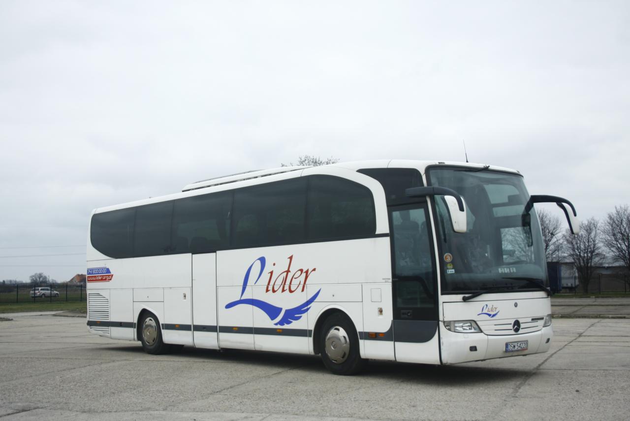 Biały autobus turystyczny marki Mercedes z logo firmy 'Lider' na boku, stojący na betonowym placu w pochmurny dzień.