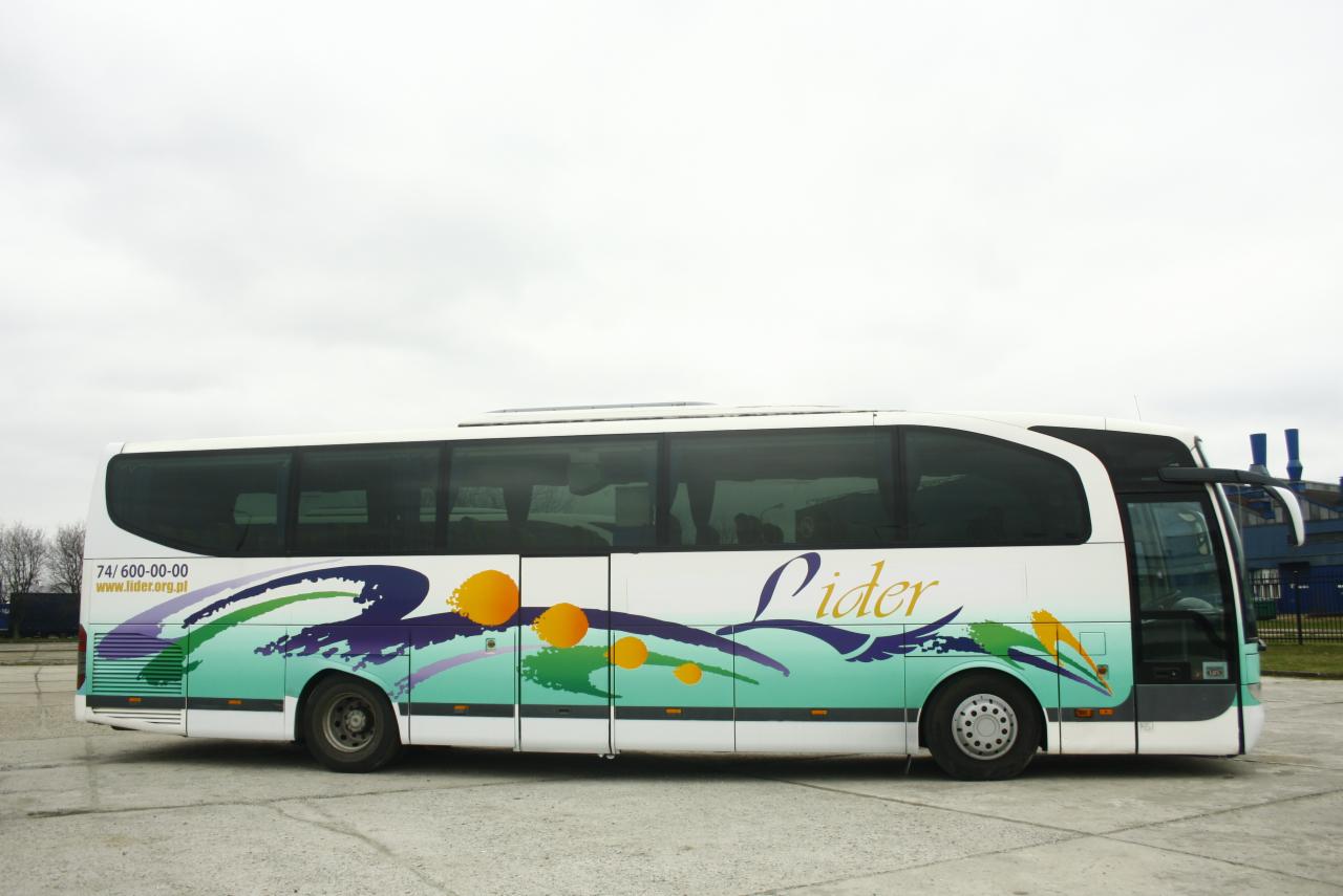 Biały autokar z kolorową grafiką i napisem 'Lider' zaparkowany na betonowym placu, widok z boku w pochmurny dzień.