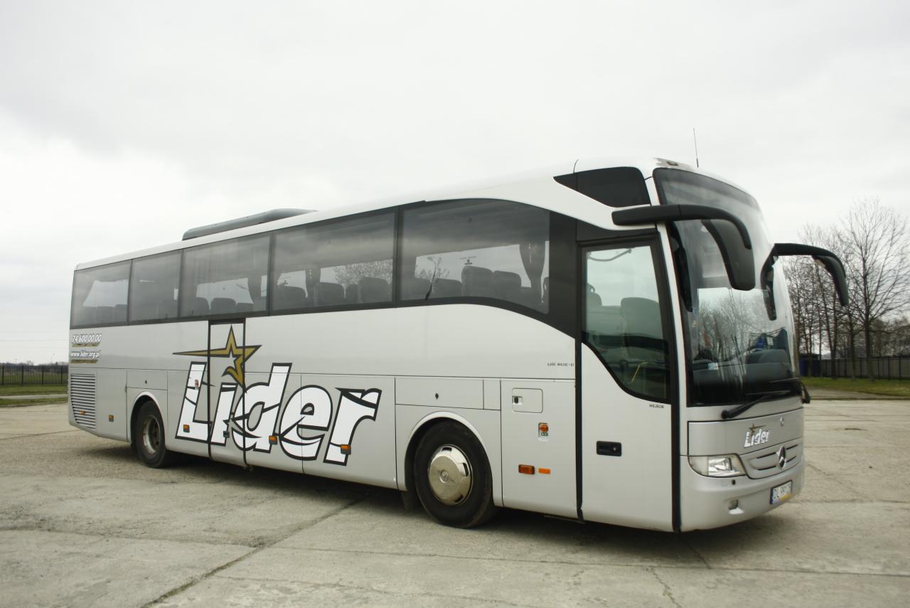 Autokar turystyczny w kolorze srebrno-białym z napisem 'Lider' i gwiazdą na boku, zaparkowany na betonowym placu w pochmurny dzień.