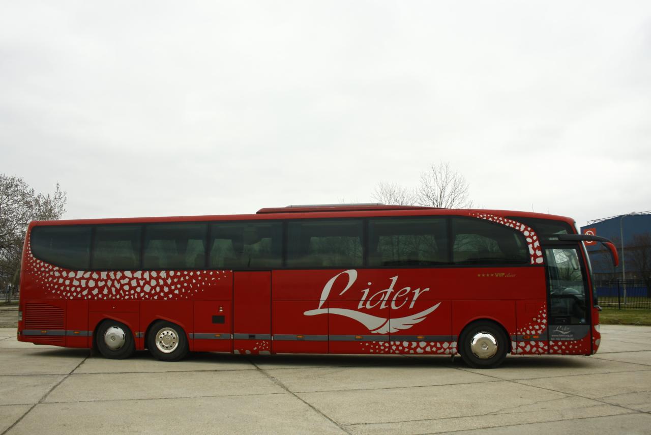 Czerwony autokar z białym napisem 'Lider' i motywem skrzydeł, oznaczony jako VIP Class, zaparkowany na betonowym placu pod pochmurnym niebem.