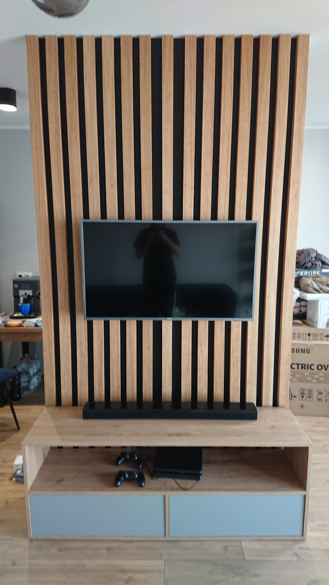 Telewizor zawieszony na ścianie z drewnianych lameli, pod nim szafka RTV z półką, na której leżą pady do konsoli.