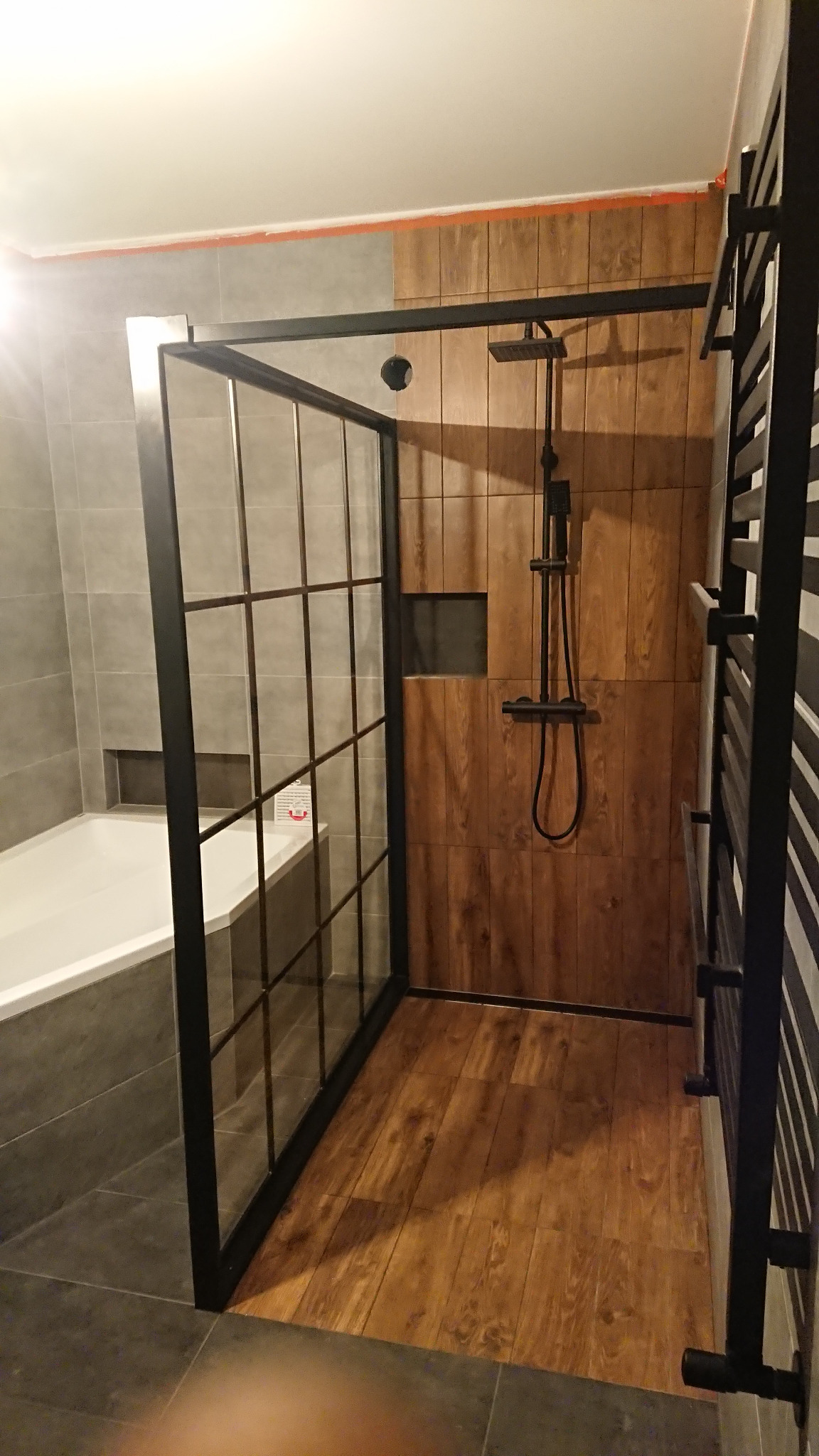 Nowoczesna kabina prysznicowa z czarną ramą i drewnopodobnymi płytkami, widoczny grzejnik drabinkowy i bateria natryskowa w czarnym macie, na podłodze drewnopodobne panele.