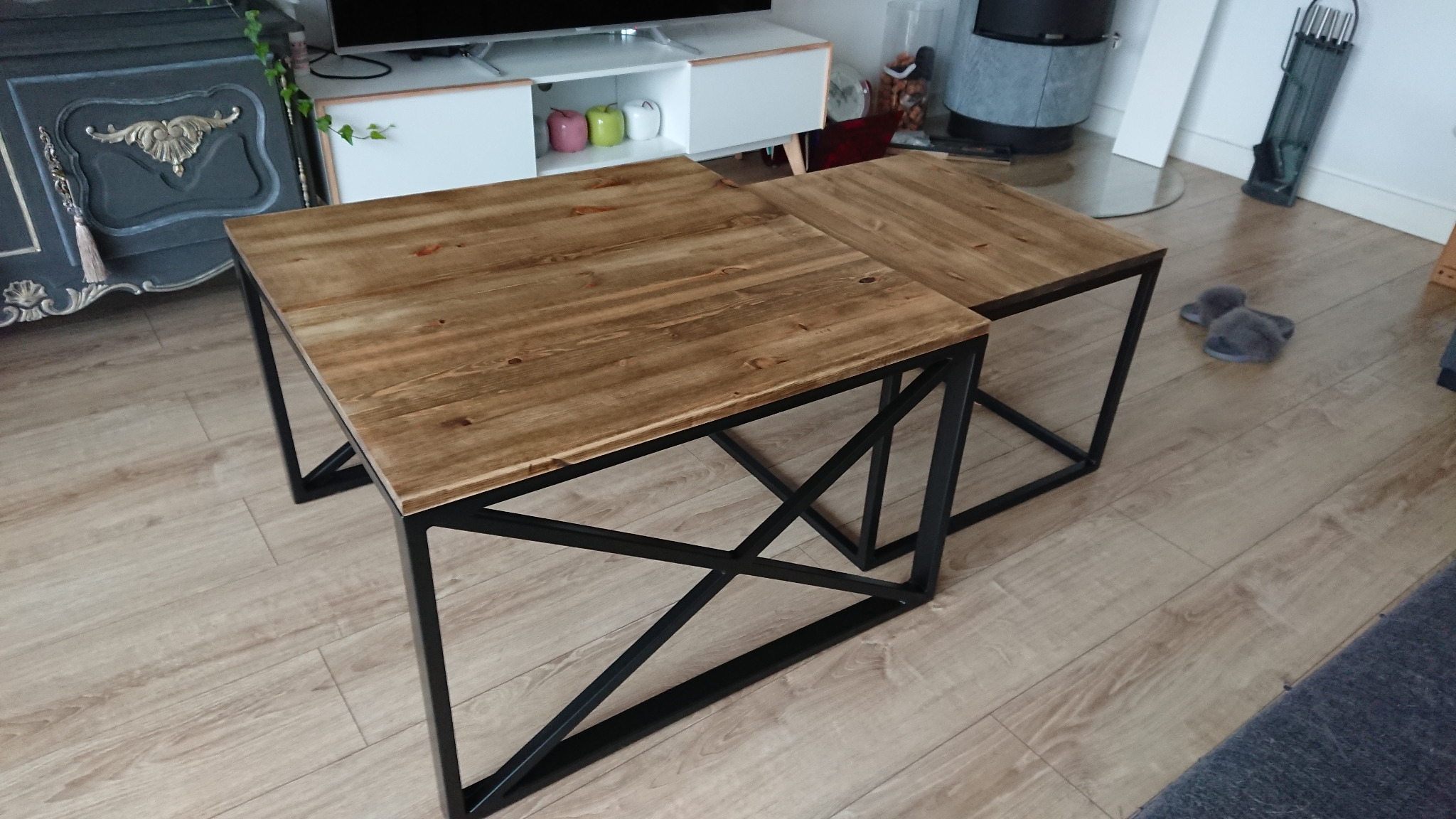 Dwa stoliki kawowe z drewnianymi blatami o ciepłym odcieniu i czarnymi metalowymi nogami o geometrycznym kształcie, ustawione na jasnej podłodze z paneli, z widocznymi elementami wystroju wnętrza...