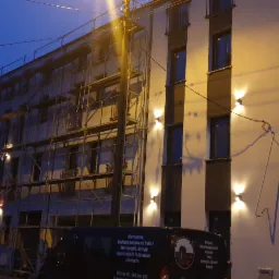 Budynek wielorodzinny w trakcie prac elewacyjnych, widoczne rusztowania i oświetlenie zewnętrzne, na pierwszym planie samochód dostawczy z logo firmy budowlanej.