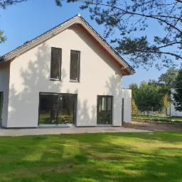 Nowoczesny, murowany dom jednorodzinny z białymi ścianami i ciemną stolarką okienną, częściowo ocieplony, na działce z zielonym trawnikiem, widoczne drzewa iglaste.