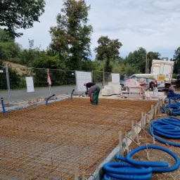 Prace zbrojarskie na budowie: dwaj pracownicy montują siatkę zbrojeniową na folii, widoczne niebieskie rury karbowane, szalunki oraz maszyny budowlane w tle.