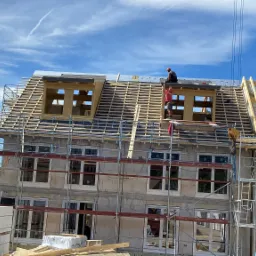 Wznoszenie dwupiętrowego budynku z pustaków z drewnianą konstrukcją dachu i oknami, widoczne rusztowania i pracownicy na różnych poziomach, w tle zielone drzewa i błękitne niebo.