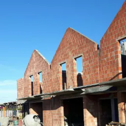 Wznoszenie ścian z czerwonej cegły na budowie szeregu domów jednorodzinnych, widoczna betoniarka i palety, błękitne niebo w tle.