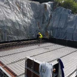 Przygotowanie zbrojenia fundamentów pod budynek na zboczu, zabezpieczonym folią, widoczny pracownik w żółtej kamizelce i kasku, elementy szalunku i siatka zbrojeniowa.