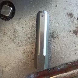 Wielokątny, metalowy element obrobiony na tokarce, leżący na brudnym, metalowym stole warsztatowym z widocznymi śladami obróbki i niebieskimi śladami po markerze.