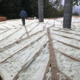 Aplikacja białej piany izolacyjnej między drewnianymi krokwiami na dachu budynku w trakcie budowy, widoczne kominy wentylacyjne i pracownik w oddali.