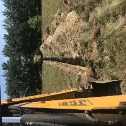 Widok z kabiny koparki JCB na wykopany wąski rów w trawiastym terenie, z pryzmami ziemi po obu stronach, w tle drzewa.