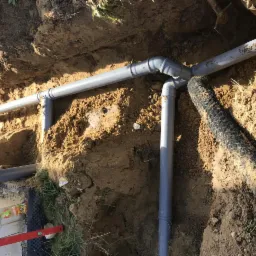 Szare rury kanalizacyjne ułożone w wykopie ziemnym, z widocznym trójnikiem i fragmentem rury drenarskiej w geowłókninie, częściowo zasypane ziemią.