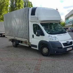 Biały bus dostawczy z szarą plandeką na parkingu z kostki brukowej, w tle budynek biurowy i inne zaparkowane pojazdy, widoczne drzewa.