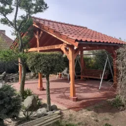 Drewniana altana z czerwoną dachówką, huśtawką i wypoczynkowymi krzesłami na kostce brukowej w ogrodzie.