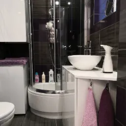 Nowoczesna łazienka z kabiną prysznicową narożną, umywalką nablatową i fioletowymi akcentami kolorystycznymi na płytkach i ręcznikach.