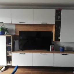 Białe meble na wymiar w salonie z drewnianym blatem, czarnymi uchwytami, telewizorem, konsolą Playstation 5, otwartą szafką z alkoholem, roślinami w niebieskich doniczkach i zdjęciem w ramce.